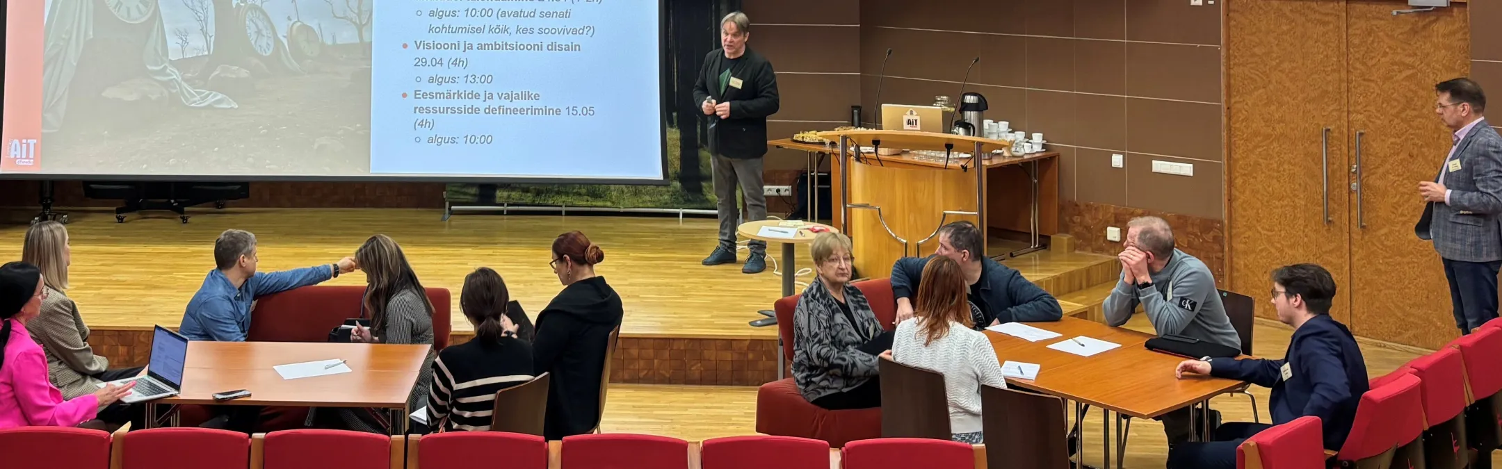 Üks inimene seisab laval ekraani ees ja teised istuvad laudkondades ning kuulavad tema ettekannet